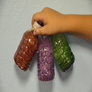 Child's hand holding three sensory bottles with glitter. One is orange, one is purple, and one is green.