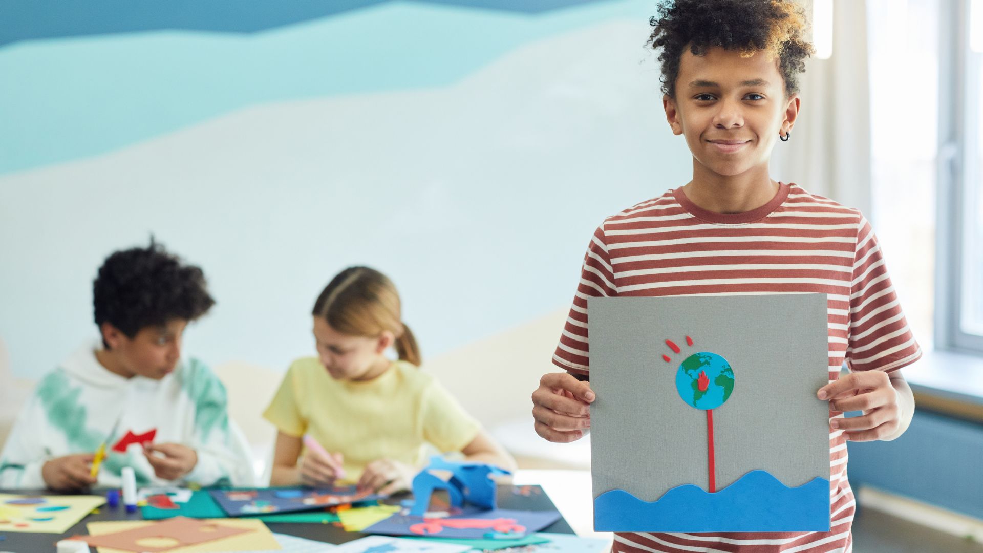 One elementary school youth in the foreground holding felt artwork. Two youth in the background are creating their felt artwork.