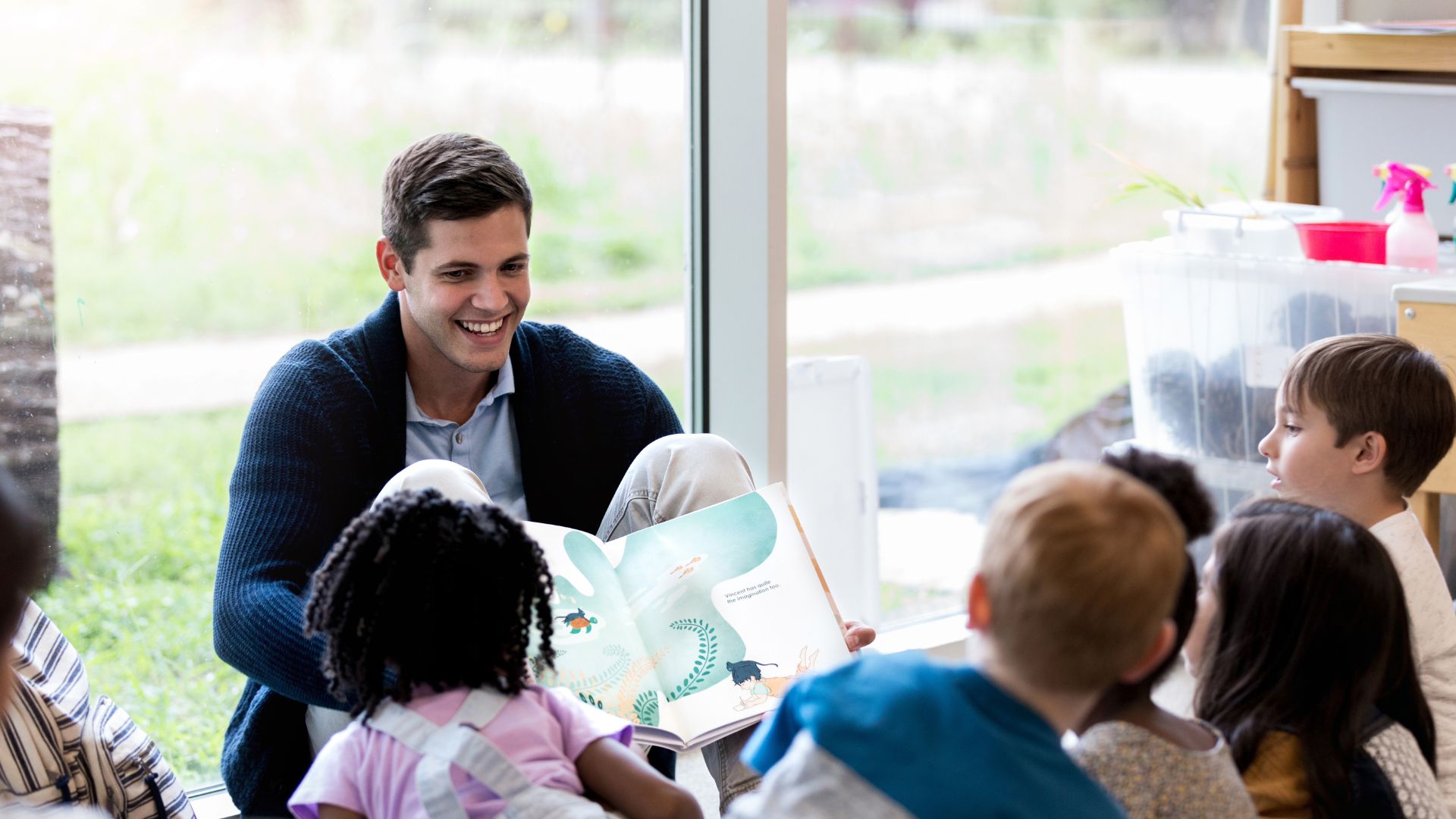 Teacher smiling while holding a picture up to a group of preschool children.