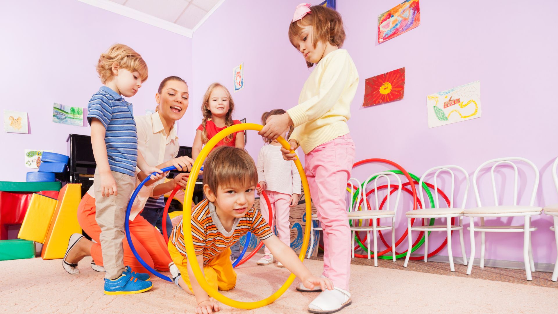 A teacher with young children are holding hula hoops above the floor for kids to crawl through