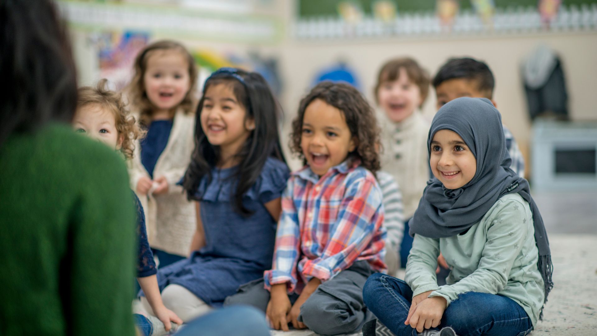 Seven preschool children sitting on the carpet looking at their teacher. The children are smiling and giving the teacher their full attention. 